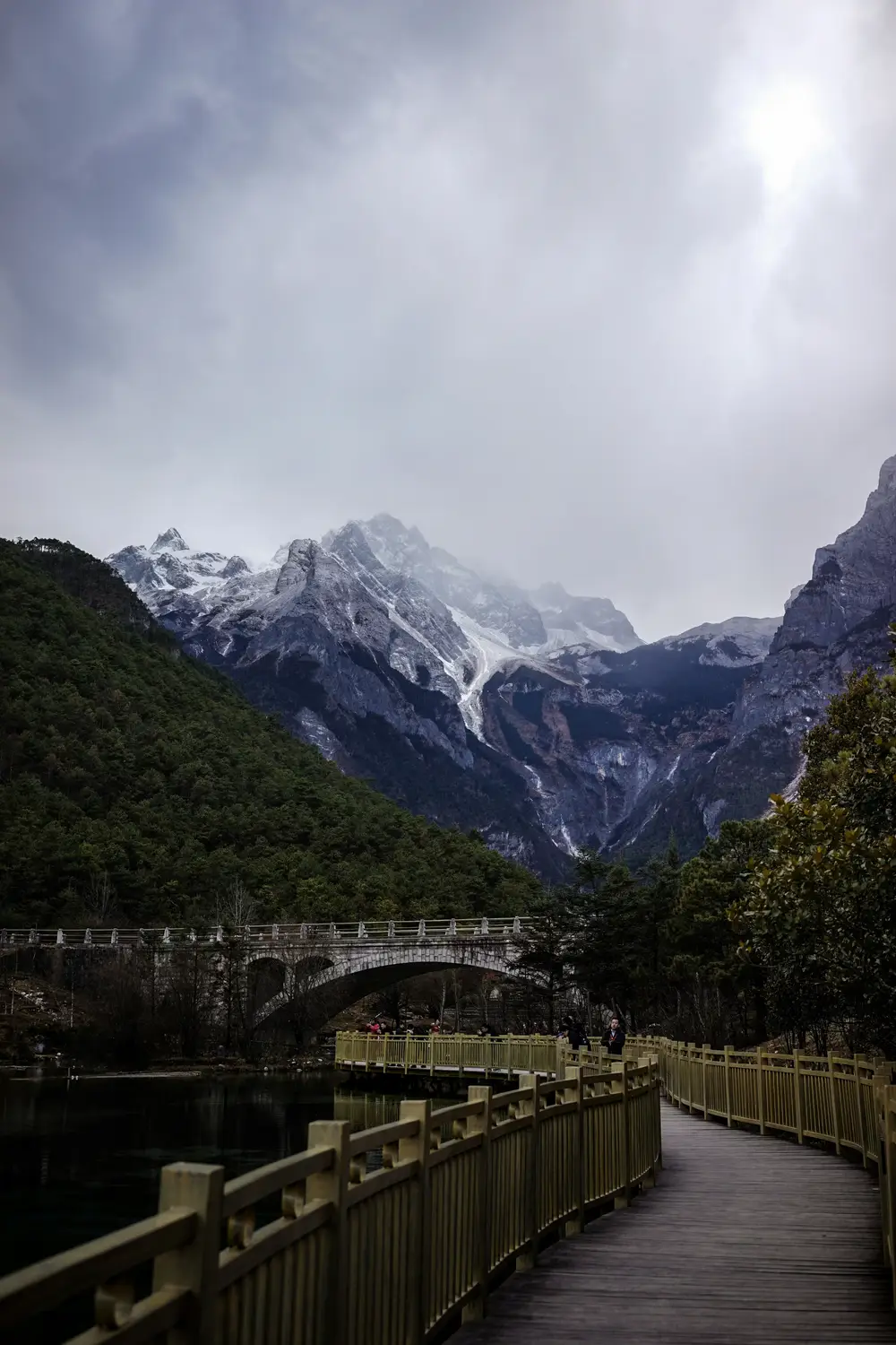 Wooden boardwalk leading to arched stone bridge with Jade Dragon Snow Mountain in background Lijiang Yunnan China