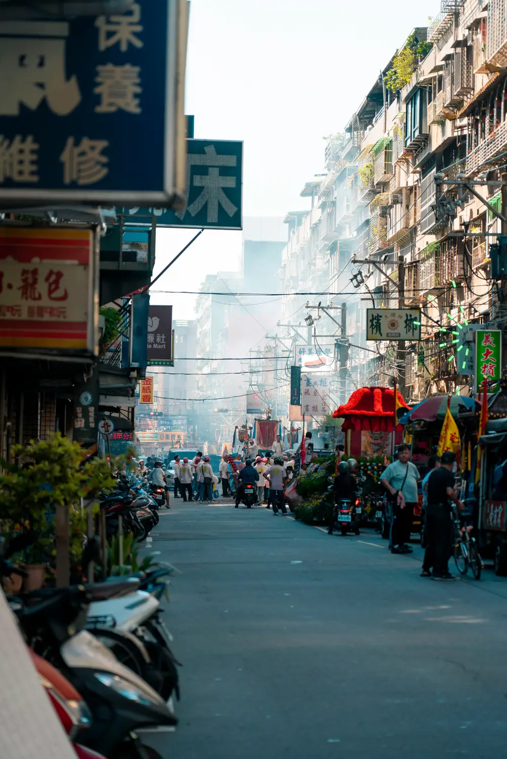 Misty Taiwan street at dawn lined with parked scooters and Chinese shop signs, with a temple procession and red palanquin in the distance