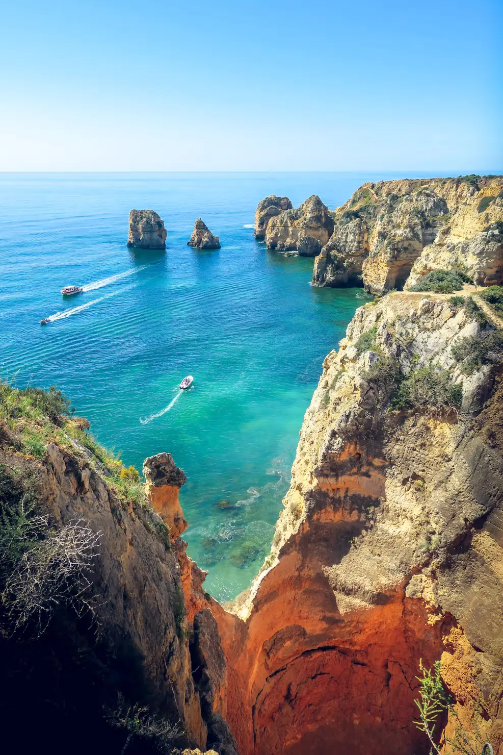 Golden coastal cliffs and sea stacks overlook clear turquoise water with small boats cruising along the shoreline.