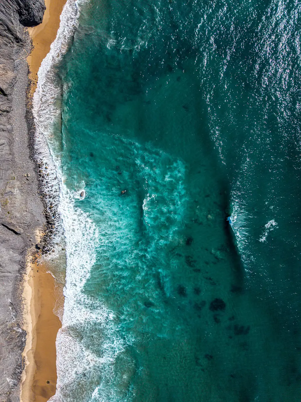 Aerial view of turquoise ocean waves breaking along a narrow sandy beach and rocky shoreline.