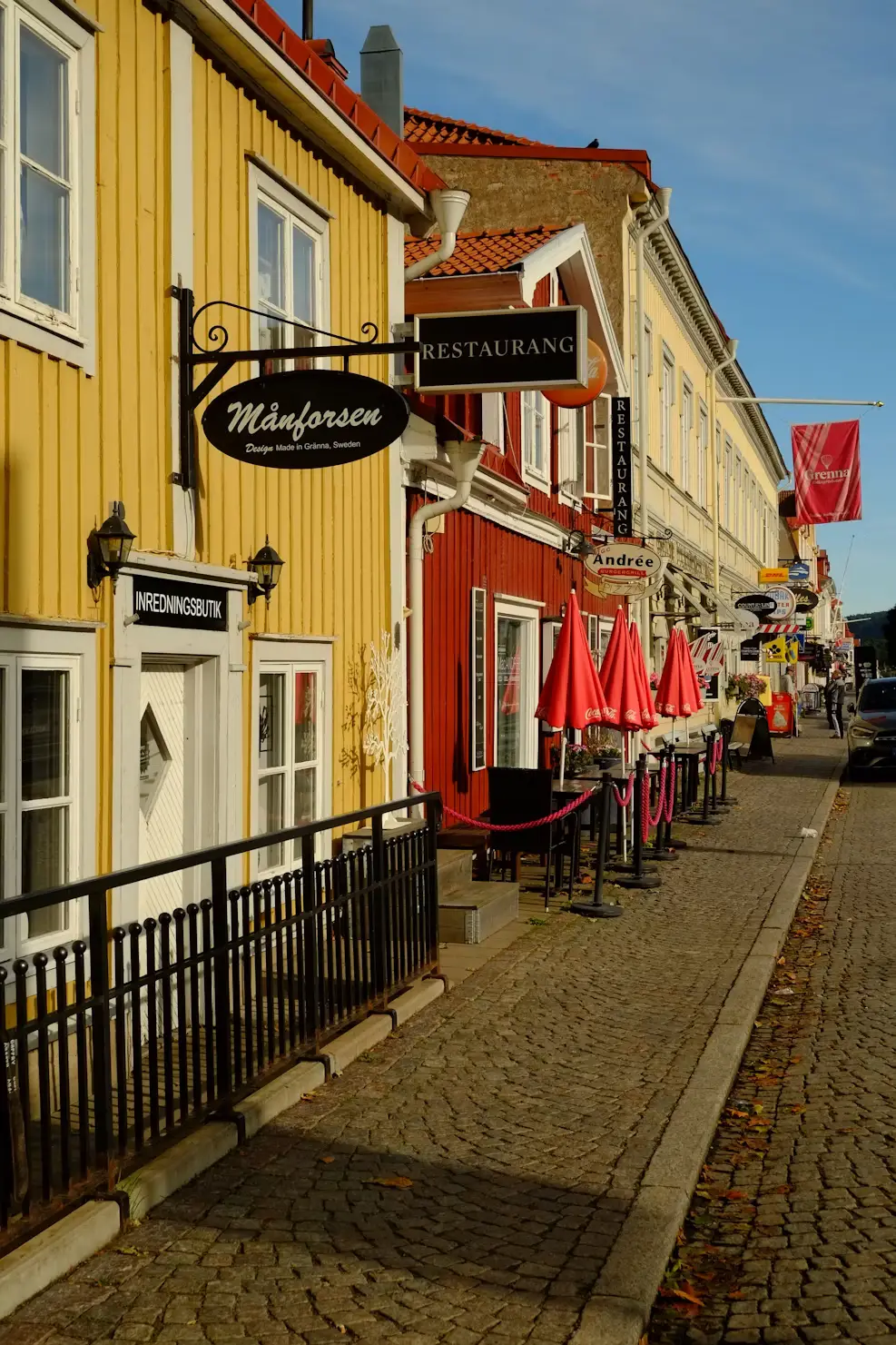Cobblestone street lined with yellow and red buildings, with outdoor café umbrellas and hanging shop signs.