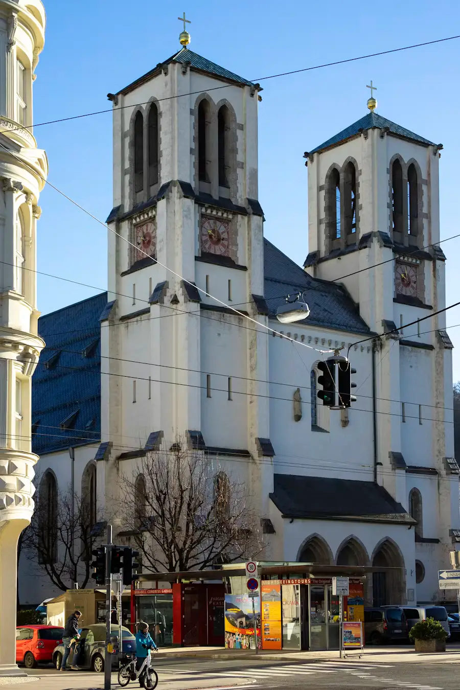 A twin-towered white church on a street corner, with a cyclist passing by and shops at street level.