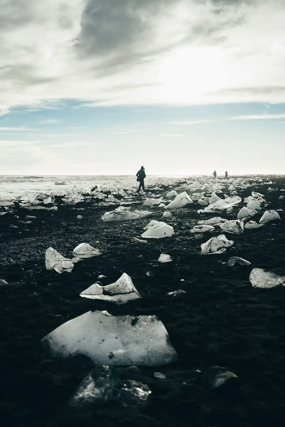 Person standing on a black sand beach scattered with glacial ice chunks under a cloudy sky, Iceland.