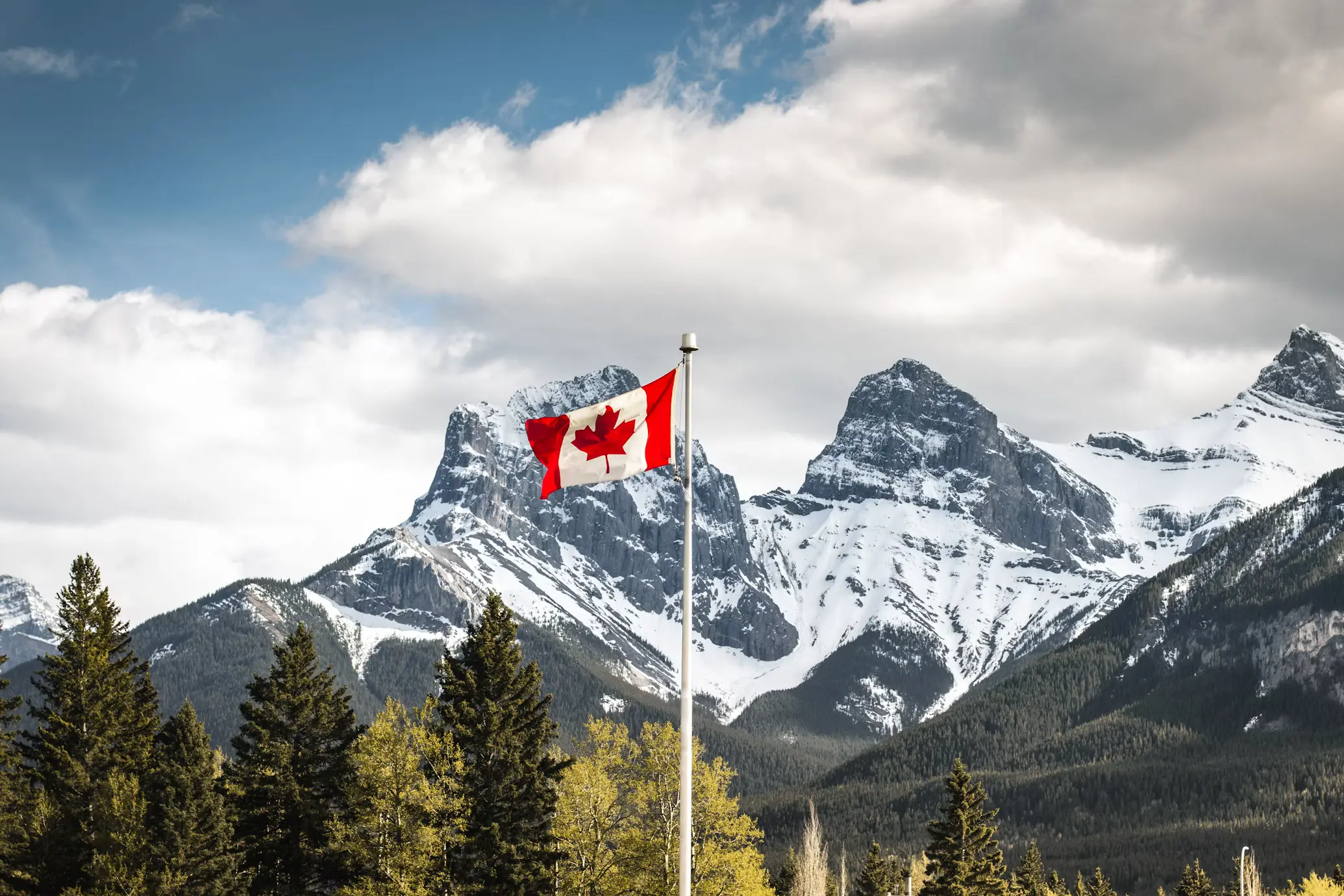 A Canadian flag flying on a pole in front of jagged, snow-dusted mountain peaks and pine forest under a cloudy sky.