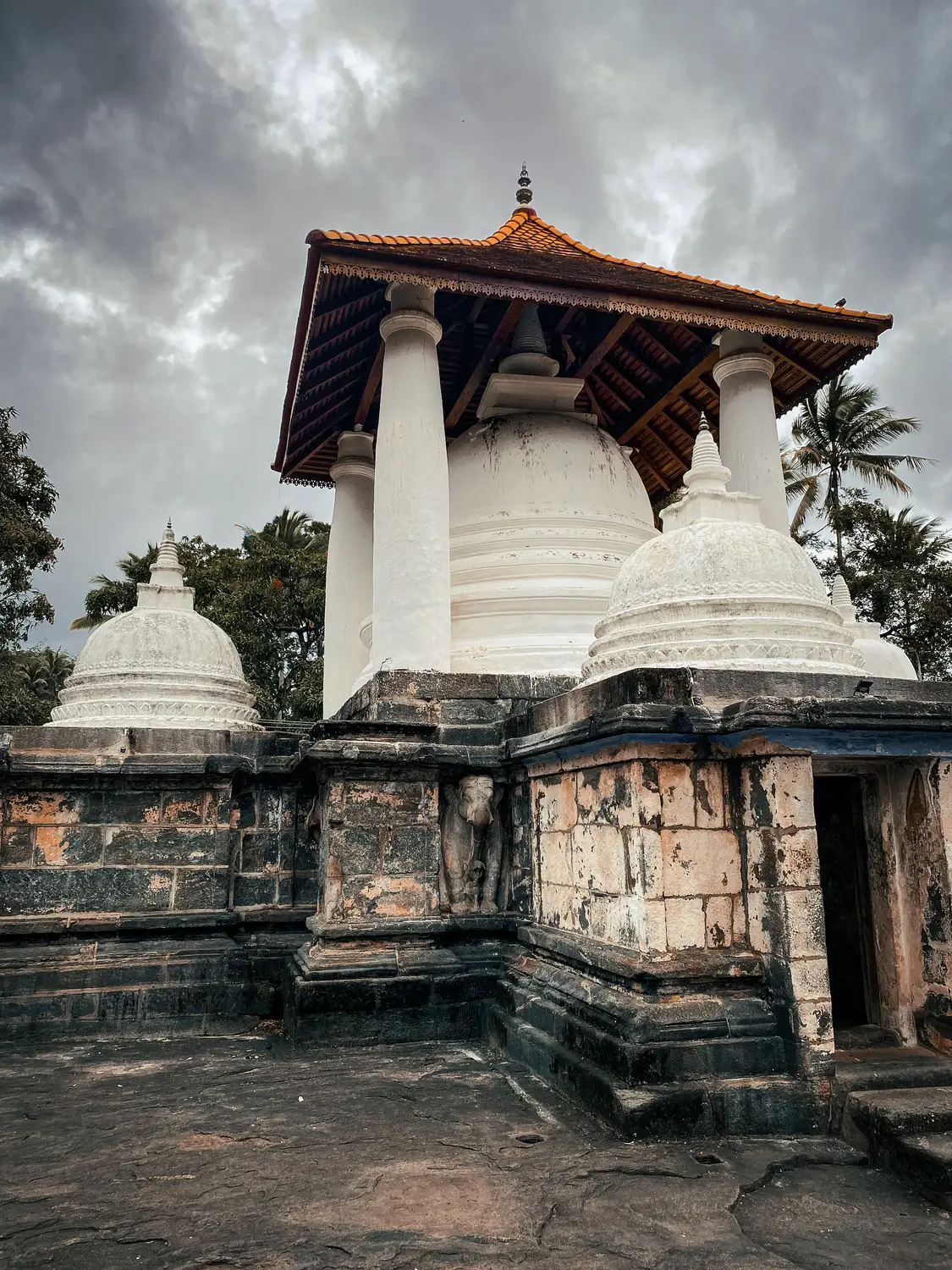 Ancient stone temple with white stupas and a tiered roof under a cloudy sky in Sri Lanka.