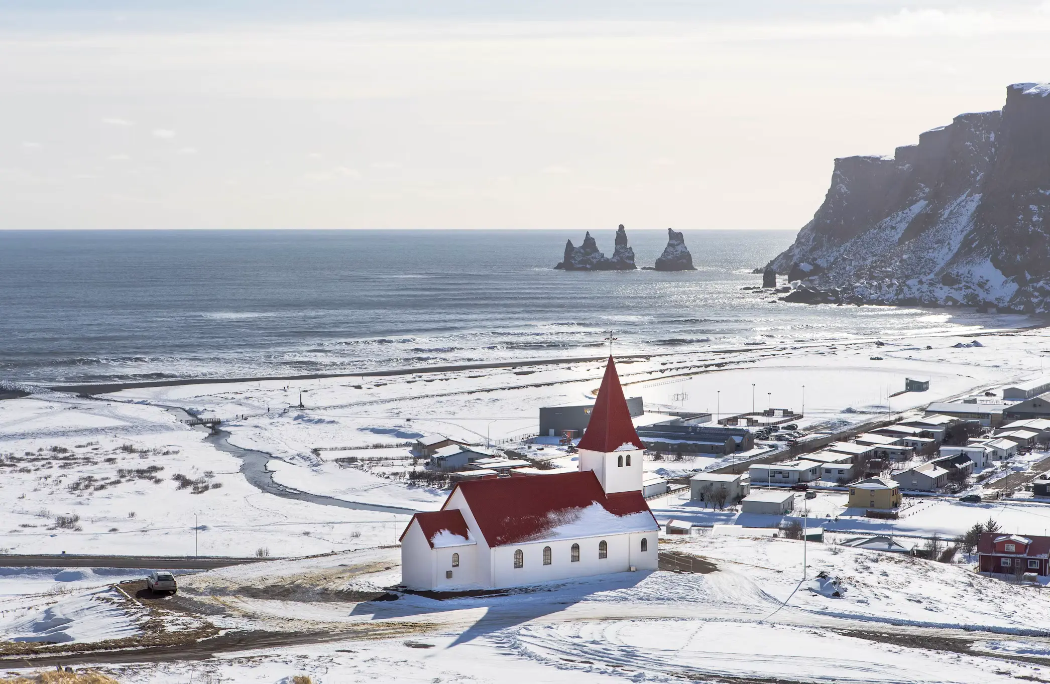 Aerial view of Vík's red-roofed church overlooking a snow-covered coastal village, with sea stacks visible in the ocean beyond, Iceland.
