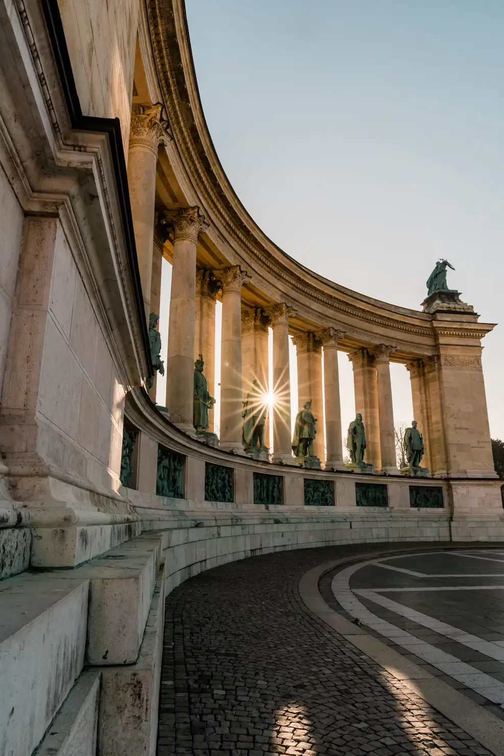 Curved colonnade with statues and sunlight shining through the columns.