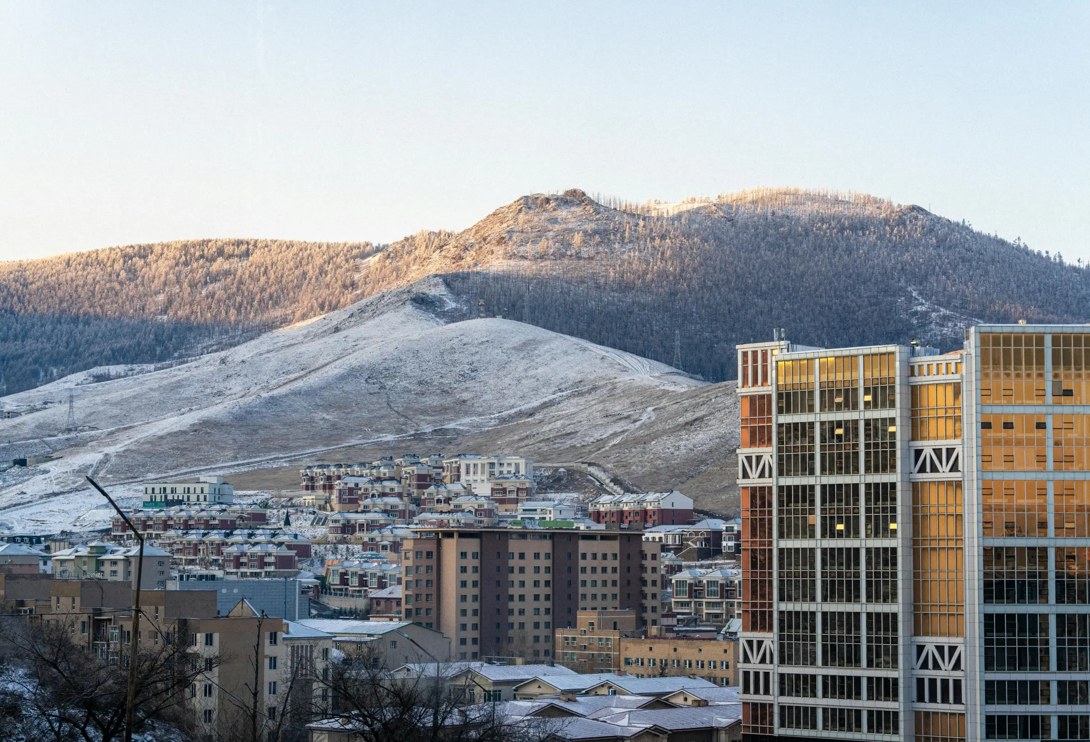 Ulaanbaatar cityscape at golden hour in winter with snow-covered Bogd Khan Mountain, residential buildings, and a modern high-rise in the foreground, Mongolia