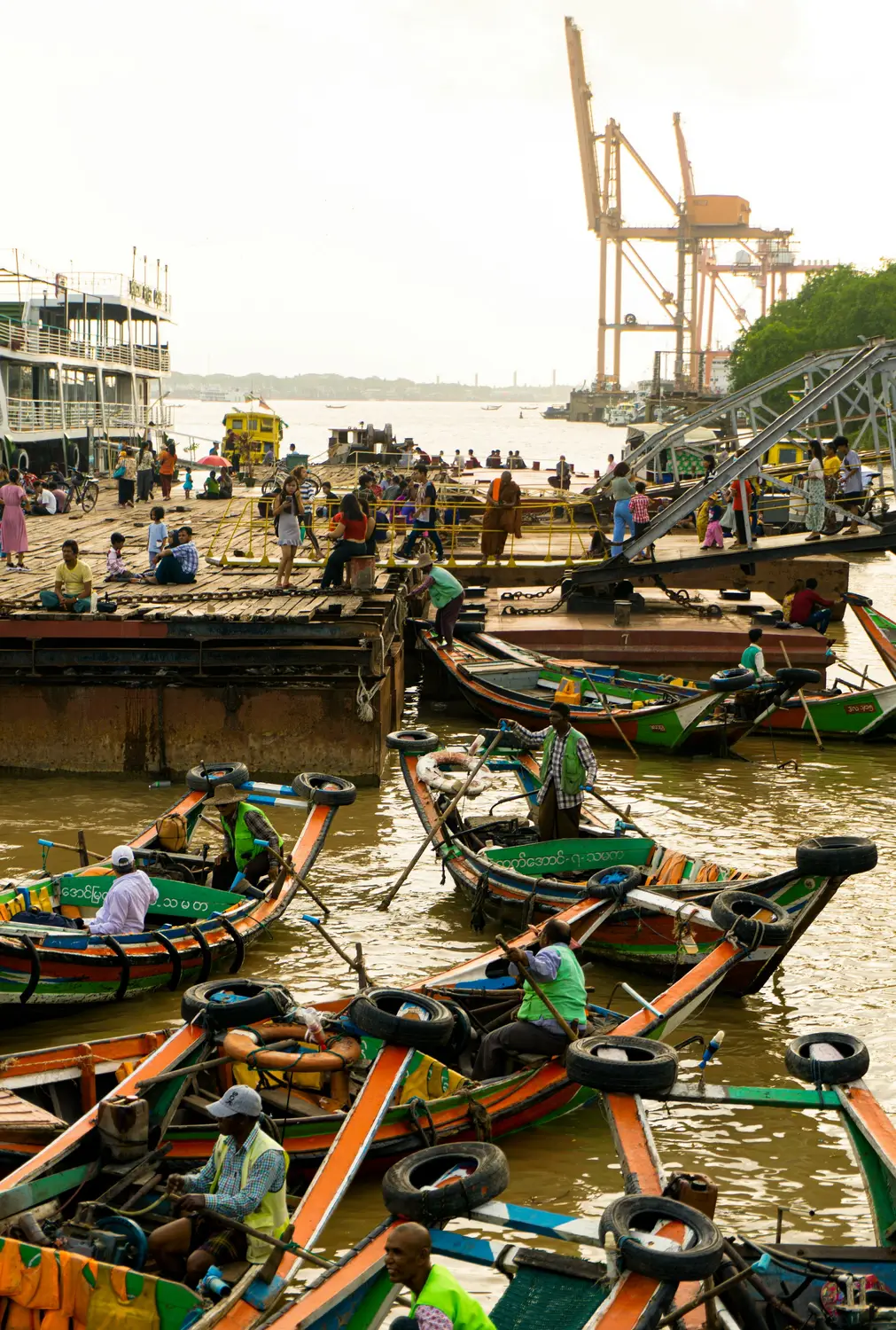 Colourful wooden taxi boats crowded at busy Yangon waterfront ferry terminal on Irrawaddy River with port cranes in background, Myanmar