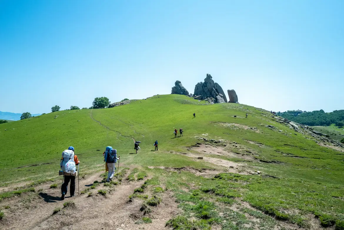 Backpackers hiking toward dramatic rock formations on a lush green Mongolian hillside