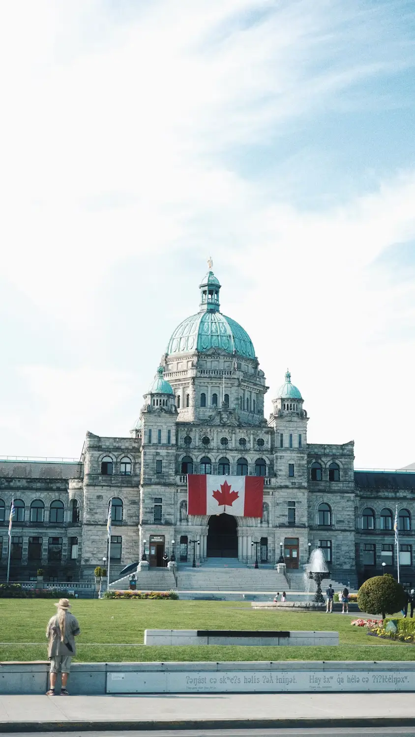 The domed British Columbia Parliament Building with a Canadian flag hanging above the entrance, seen across a lawn.