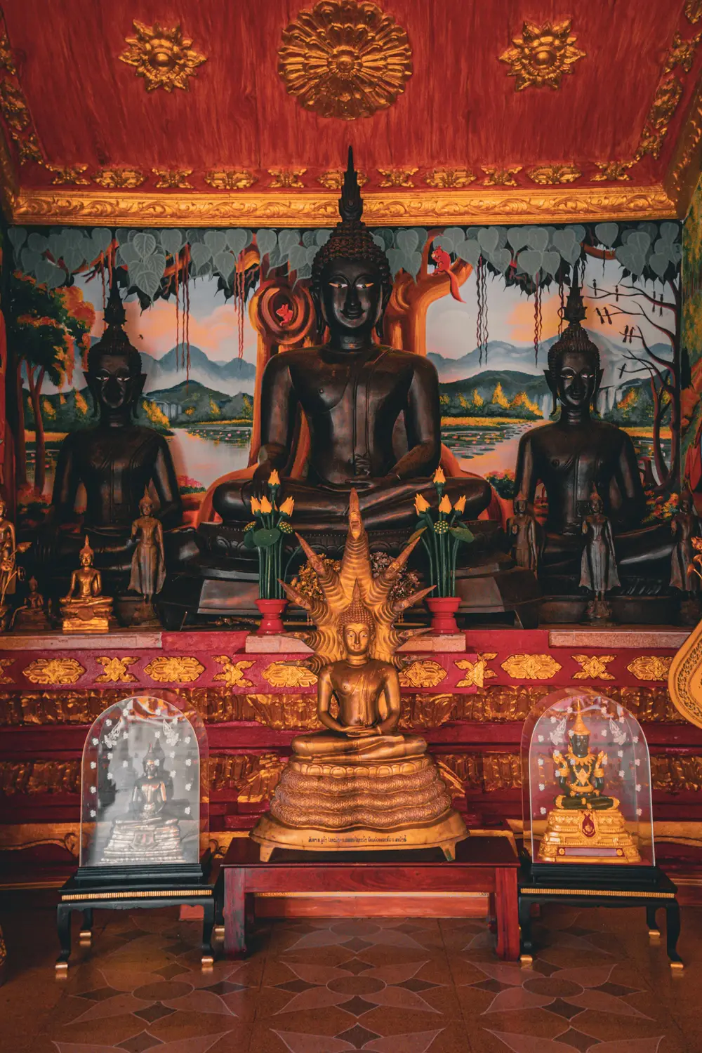 Row of dark Buddha statues against a red and gold decorated altar inside a Buddhist temple in Laos.