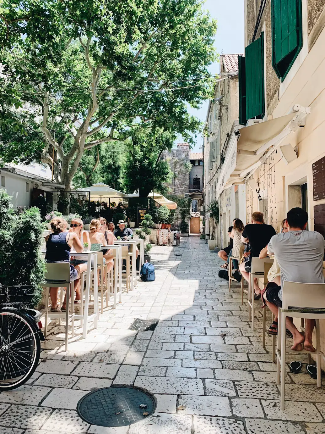 Outdoor café tables lining a shaded cobblestone alley with people seated and a bicycle parked along the wall.