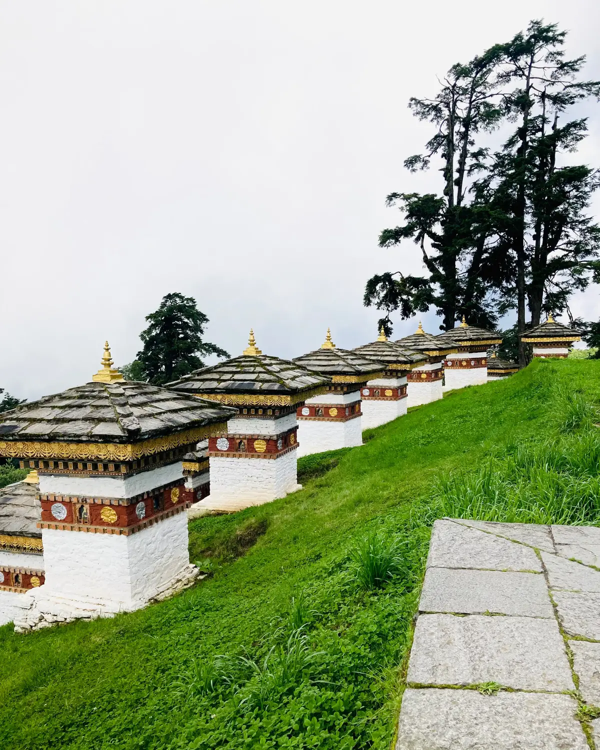 White memorial chortens lined along a grassy hillside with a stone pathway and tall trees in Bhutan.