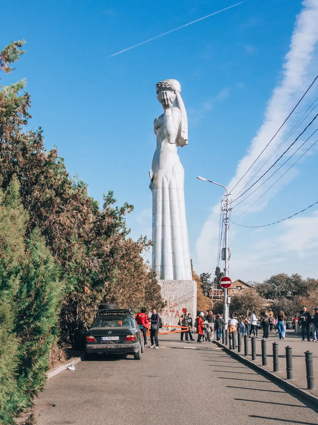 A tall stone statue beside a road, with trees, power lines, and pedestrians under a bright blue sky.