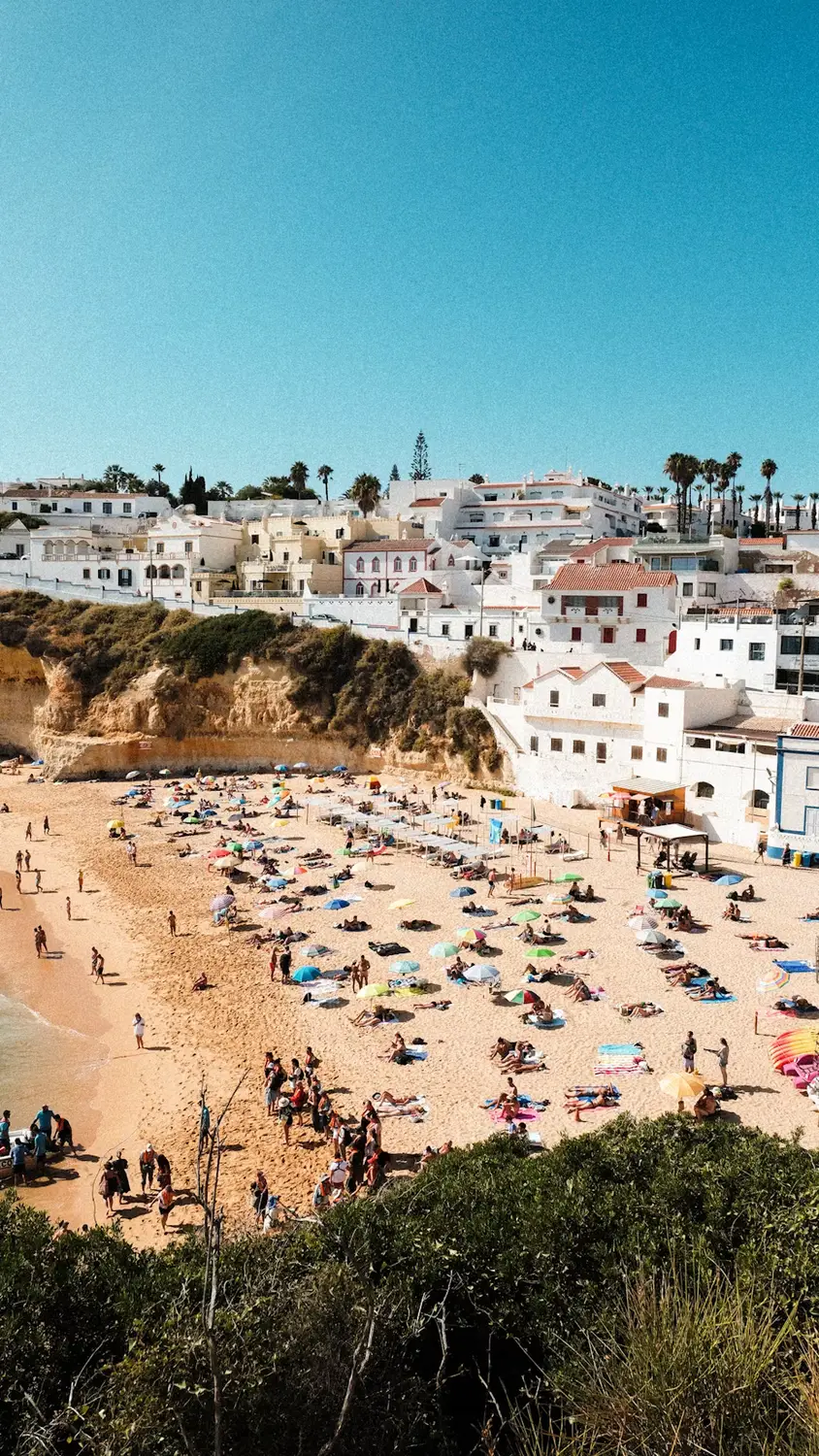A crowded sandy beach below white cliffside houses under a clear blue sky.