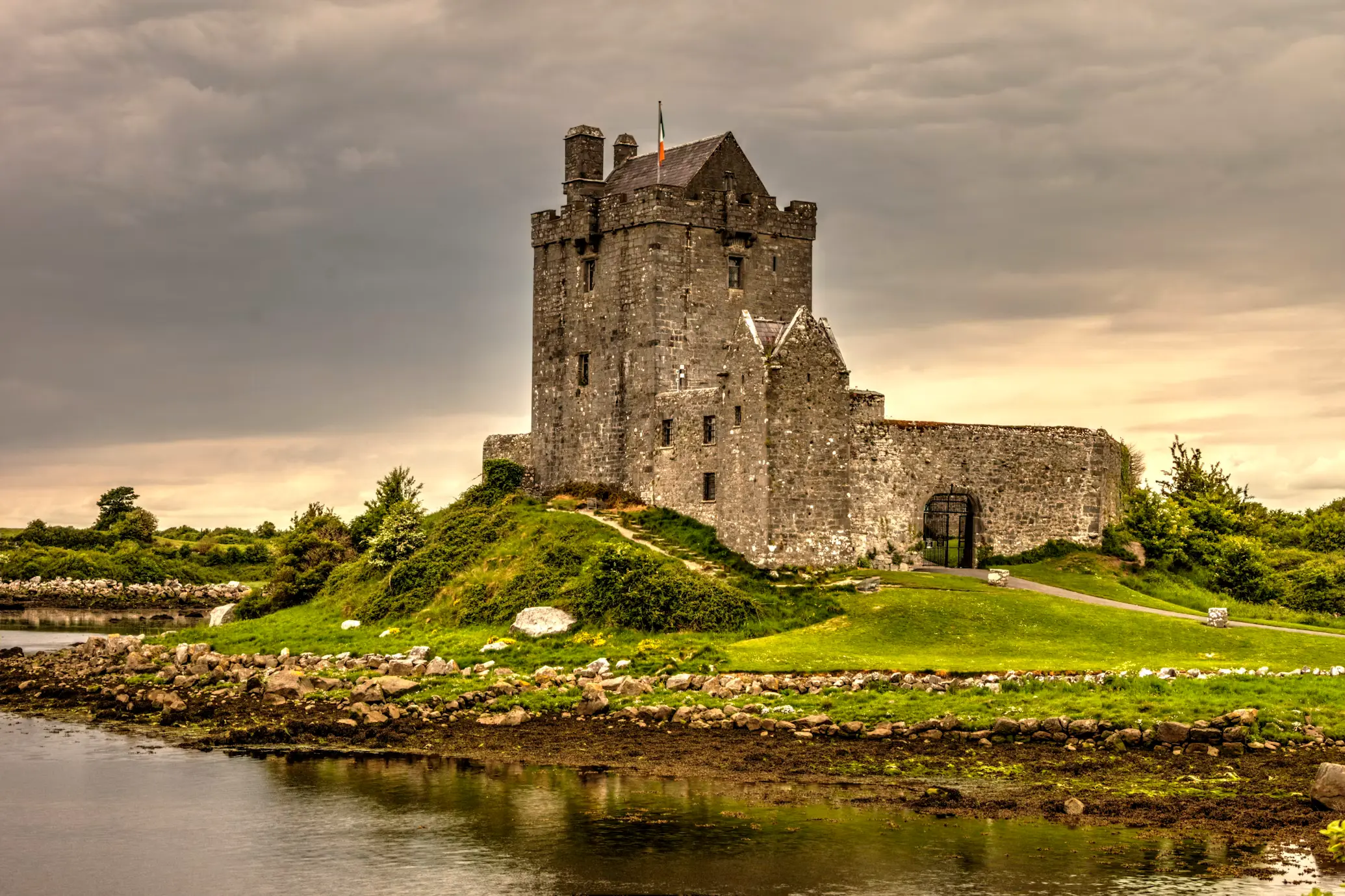 Stone tower castle beside a calm lake, surrounded by green fields under a moody sky, Ireland.