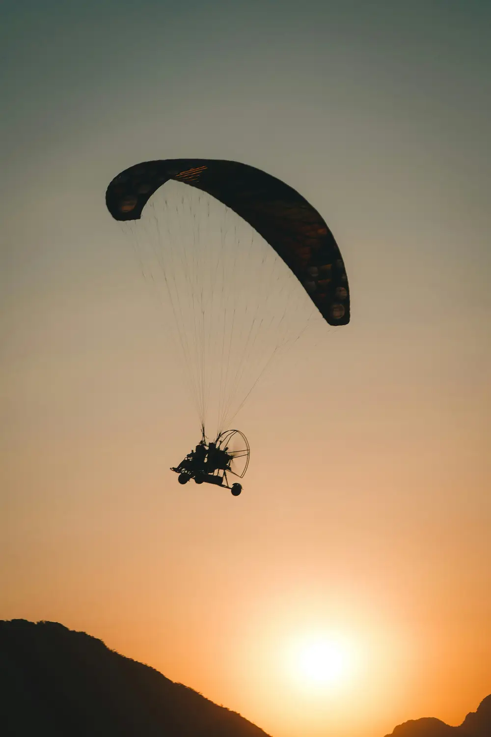 Silhouette of a powered paraglider flying against a glowing orange sunset sky over mountains in Laos.