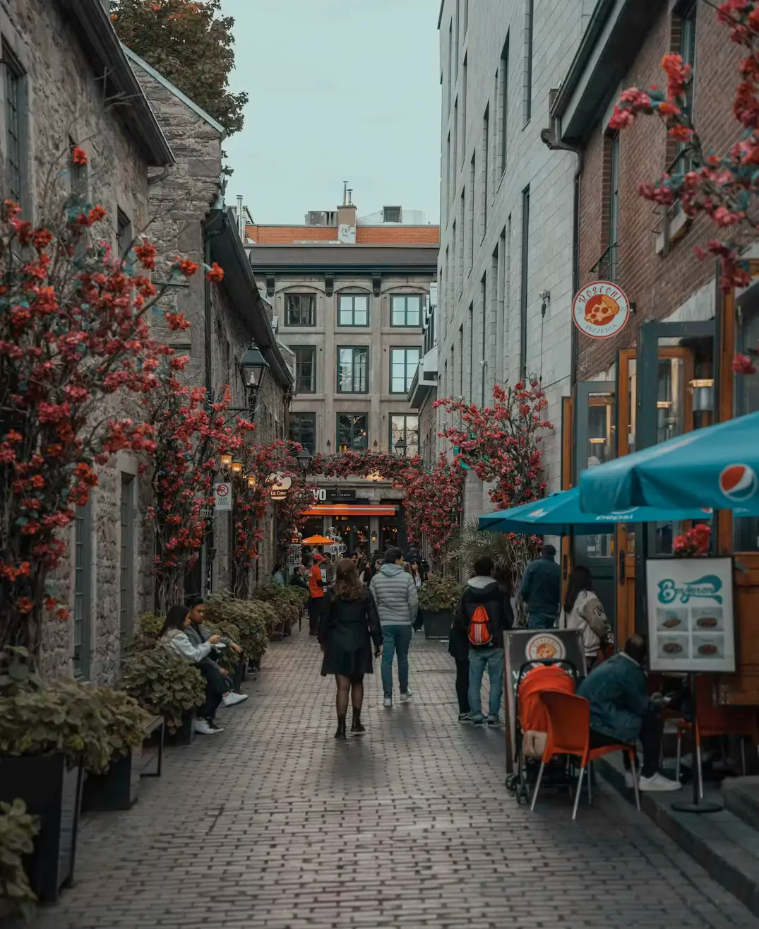 A cobblestone alley lined with flower-draped buildings, outdoor cafes, and pedestrians strolling past.