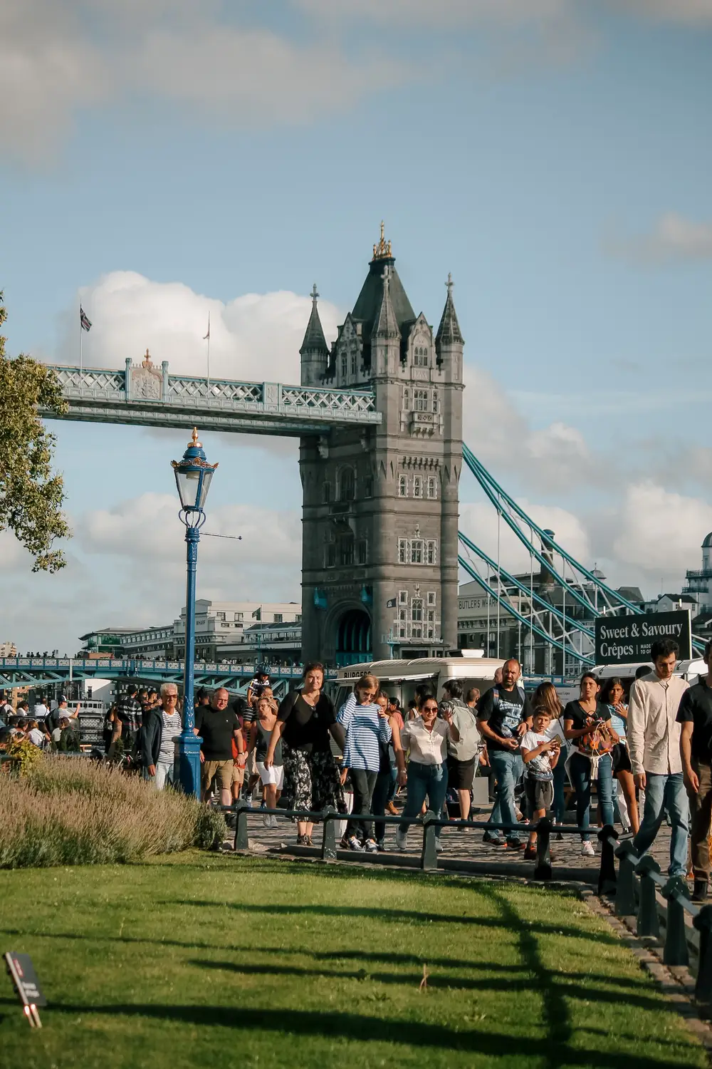 Crowds strolling along a sunny park path beside a Victorian lamppost, with Tower Bridge's stone tower rising behind them.