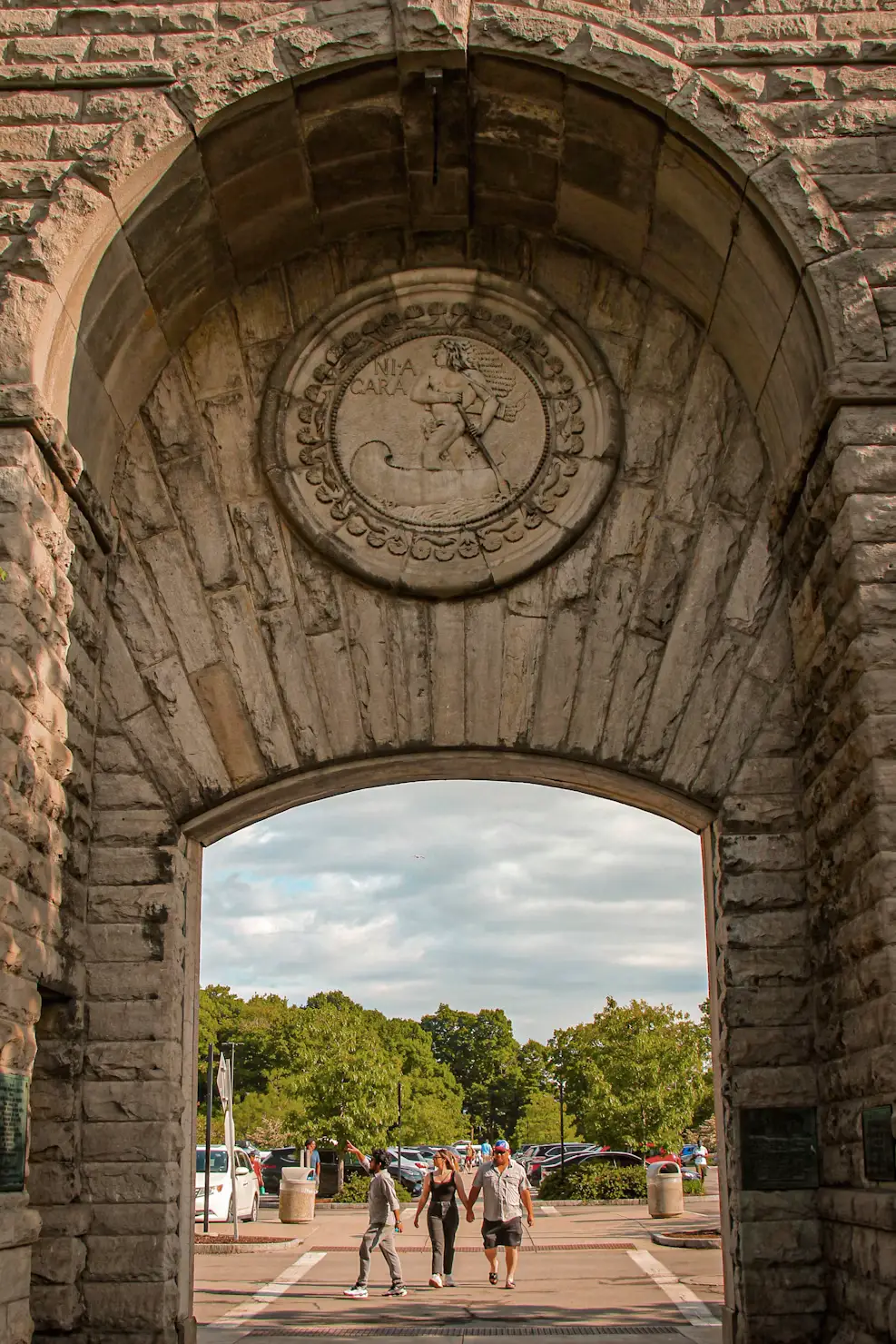A stone archway with a carved clock face, with people strolling through toward a park beyond.
