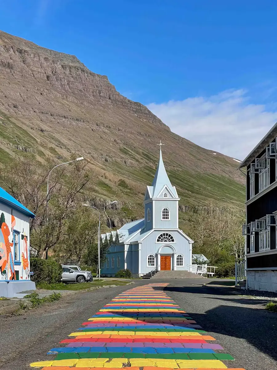 A white church at the end of a street painted with rainbow-colored crosswalk stripes, mountains rising behind, Iceland.