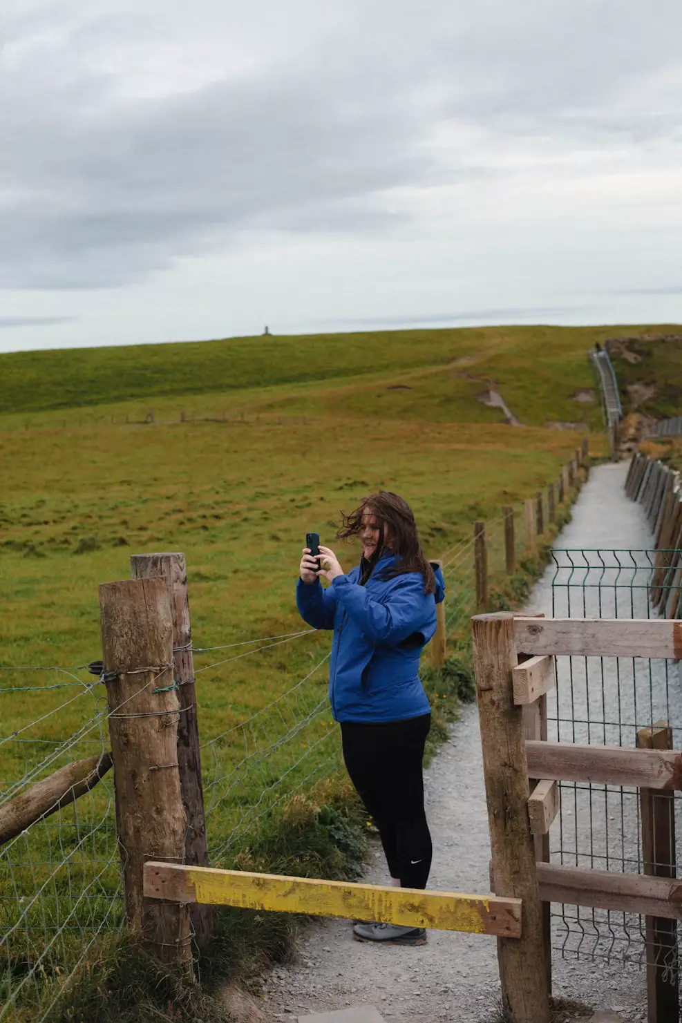 Person photographing a coastal path along a green grassy cliff with a wooden fence, Ireland.