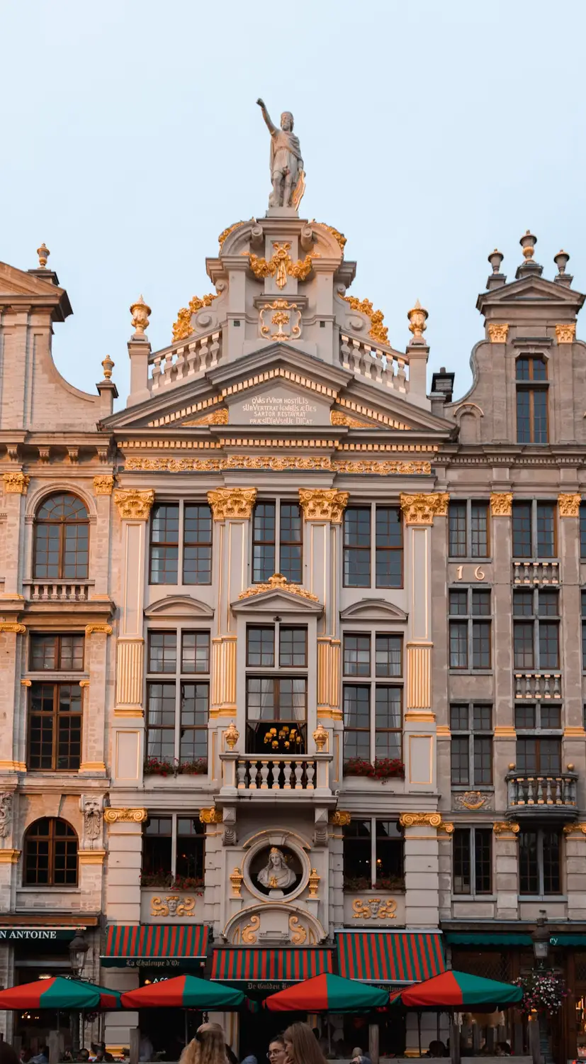 Ornate gilded facade of a Grand Place guild house in Brussels, topped with a statue, glowing in evening light with striped café awnings below.
