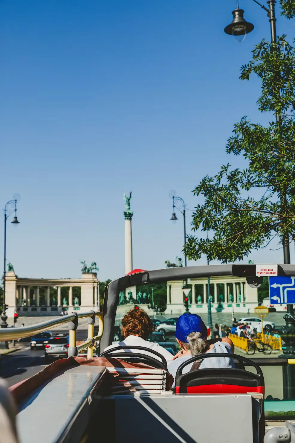 View from an open-top bus toward a grand city square with a tall column monument and historic colonnades.