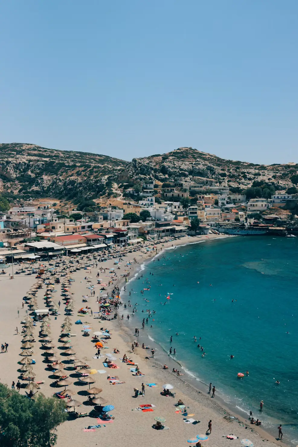 Aerial view of a Greek coastal town on a hillside, overlooking a sandy beach with umbrellas and turquoise water.