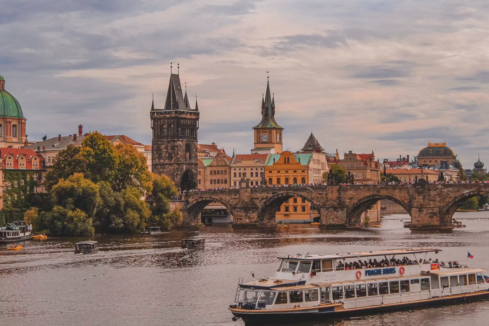 Tour boats on the Vltava River with Charles Bridge and its Gothic towers in the background under an overcast sky