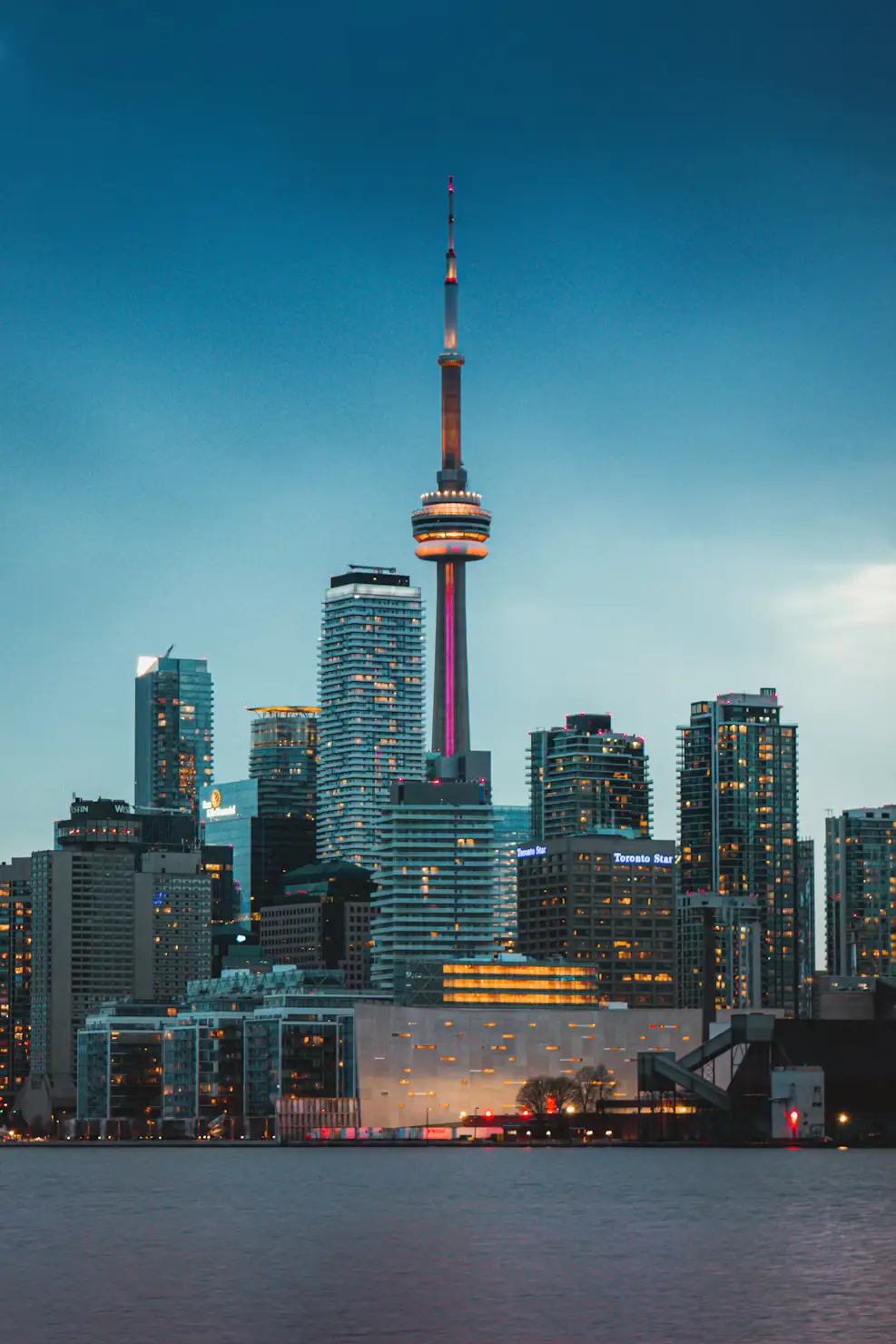 Toronto's skyline at dusk, with the CN Tower illuminated and lit skyscrapers reflecting over the waterfront.