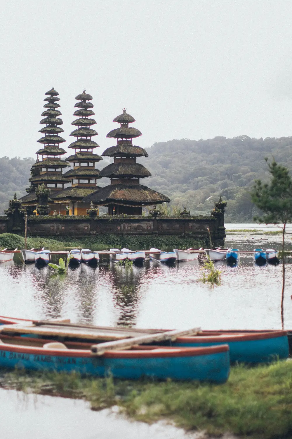 Traditional tiered meru towers of a lake temple reflected in still water, with wooden boats moored in the foreground in Bali, Indonesia.