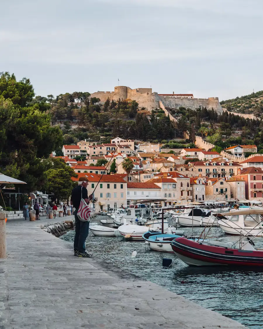 Waterfront promenade in a coastal town with a hilltop fortress overlooking red-roofed buildings and boats in the harbor