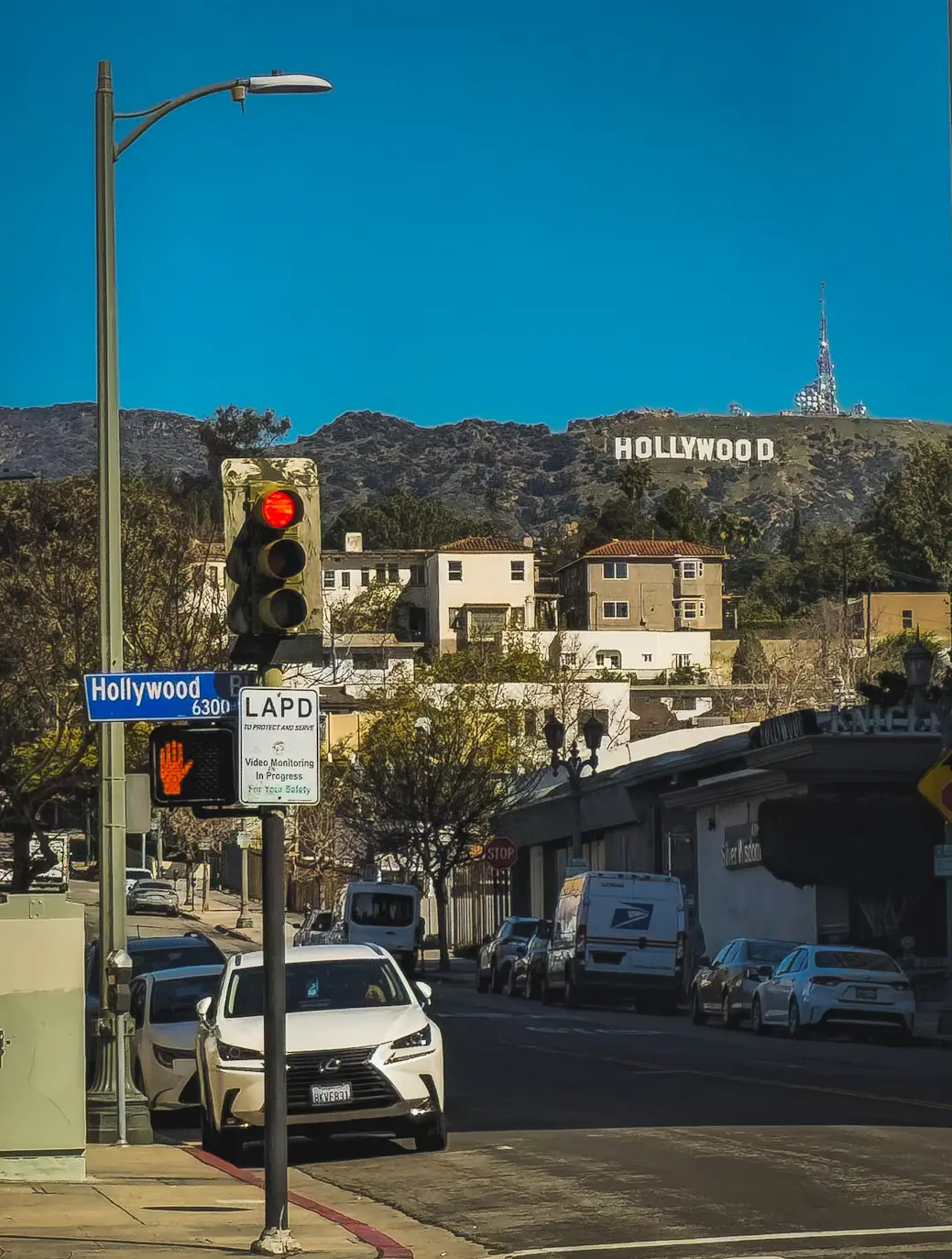 The Hollywood Sign on the hillside above a sunny Los Angeles street with cars and a traffic signal in the foreground.