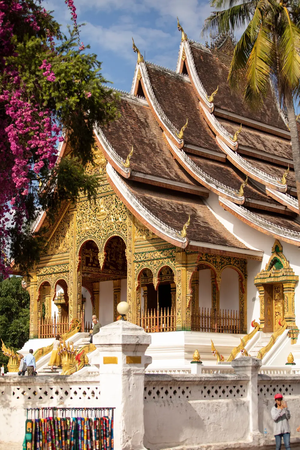 Ornate gold-decorated Buddhist temple with a sweeping tiered roof framed by pink flowers in Luang Prabang, Laos.