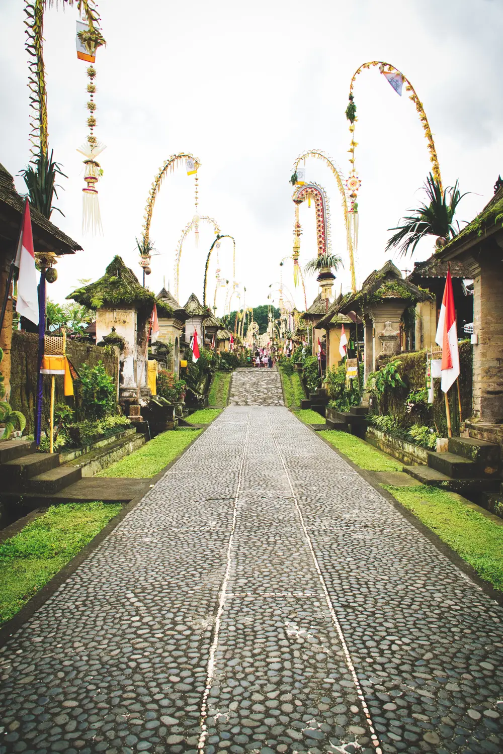 Traditional Balinese village street lined with decorative penjor bamboo poles and flags along a stone-paved road in Bali, Indonesia.