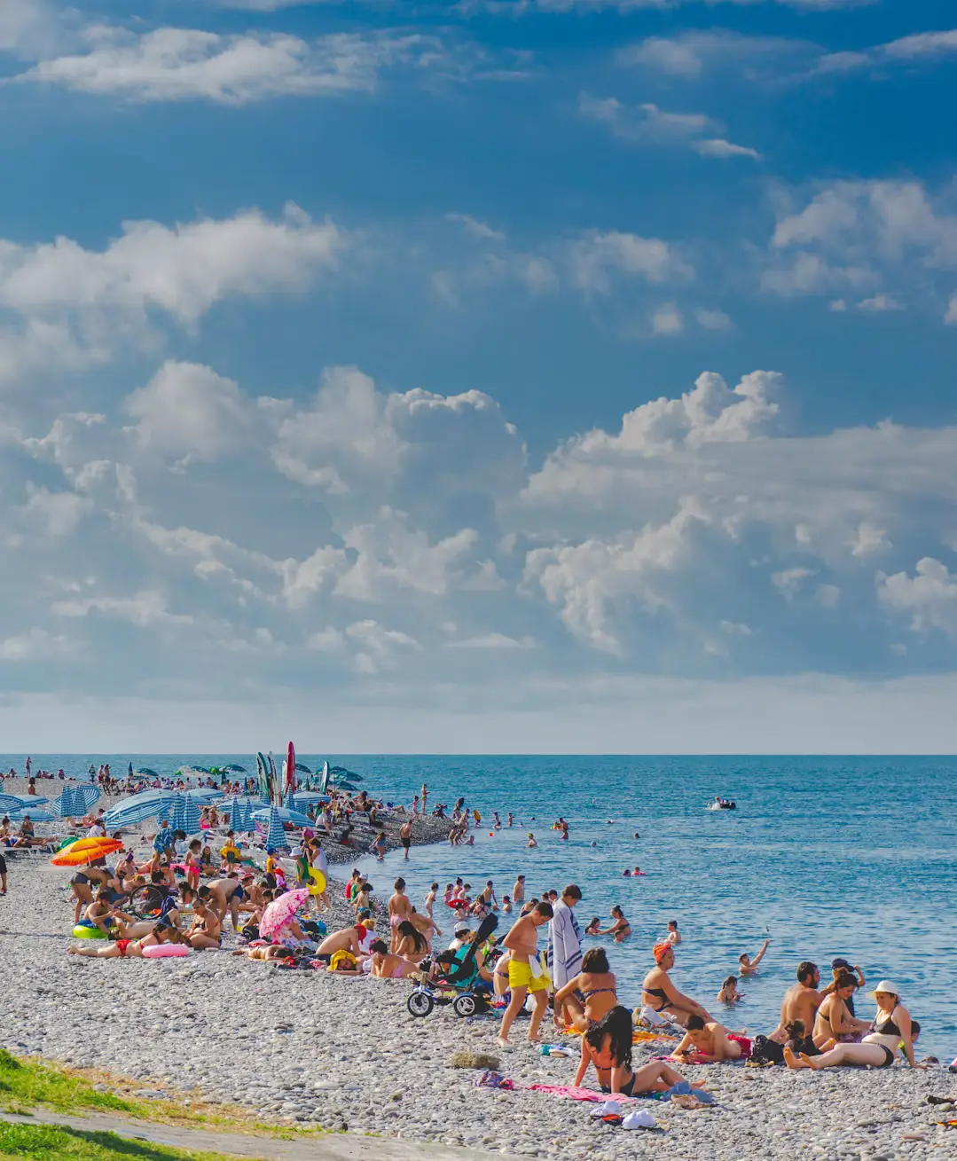 Crowded pebble beach with people swimming and relaxing under umbrellas beneath a dramatic cloudy sky.
