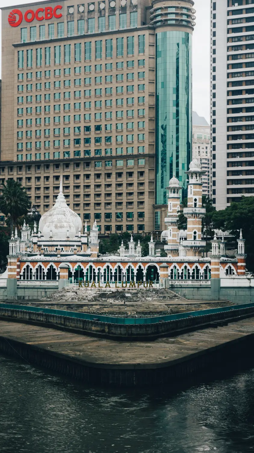 Masjid Jamek mosque with white domes and minarets surrounded by Kuala Lumpur city skyscrapers and river in Malaysia