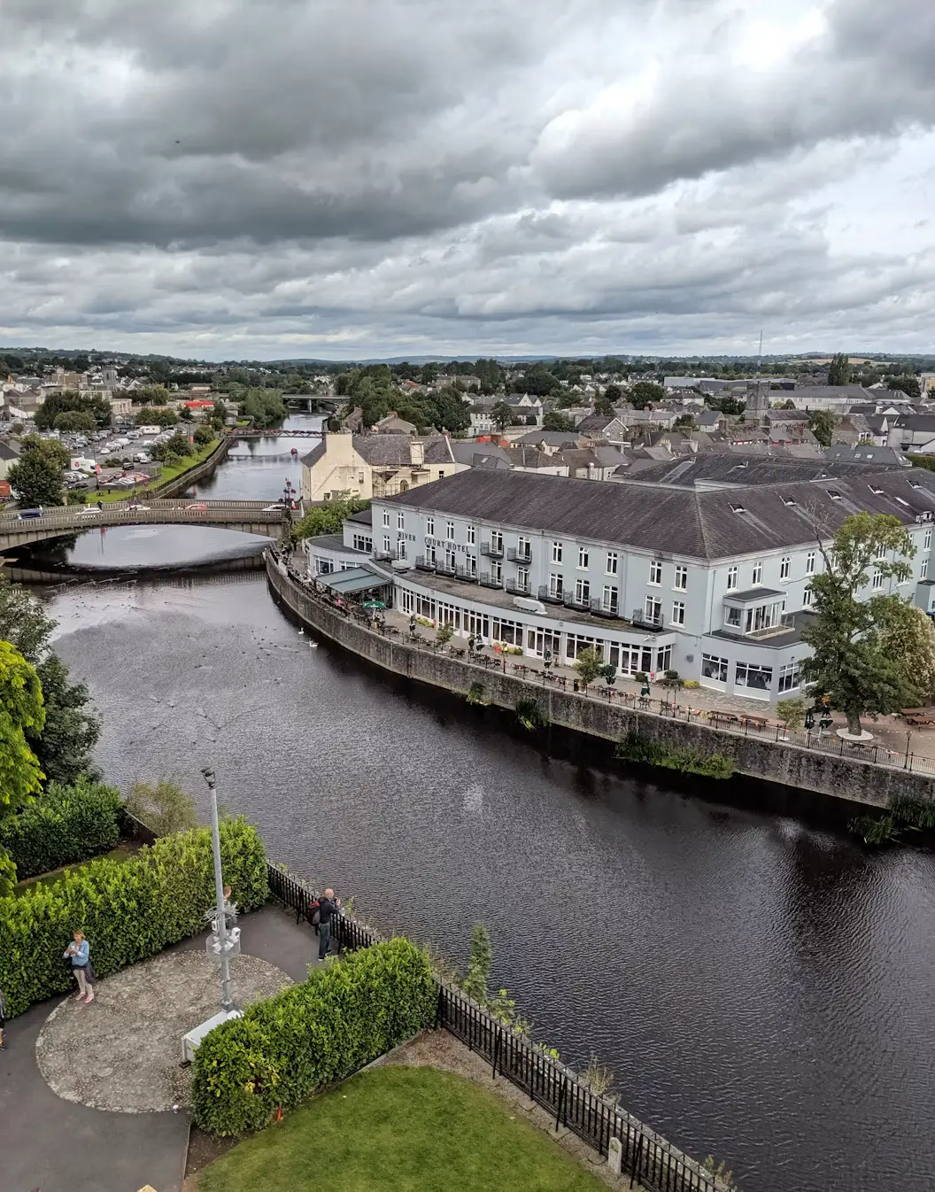 Aerial view of a river bending through a small town, lined with white buildings and a bridge, under a cloudy sky, Ireland."