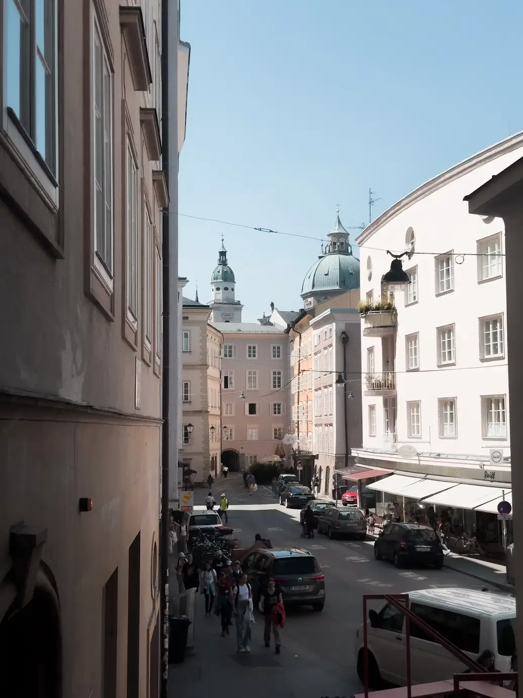A narrow street between historic buildings, with domed towers, parked cars, and pedestrians walking along the sidewalk.
