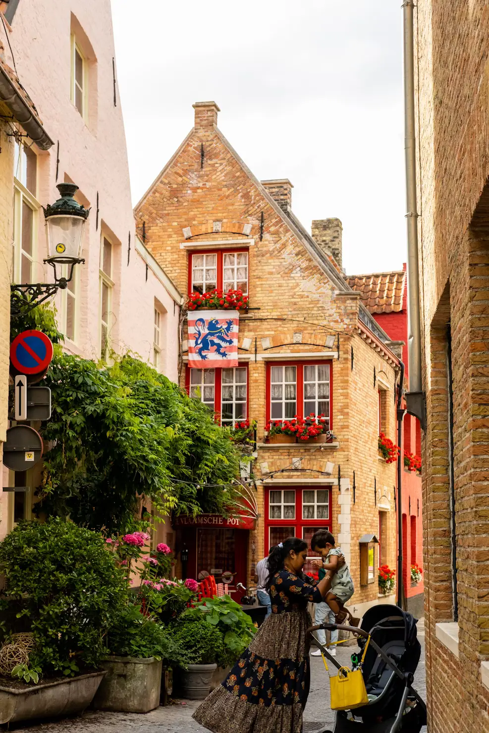 A woman and child in a narrow flower-lined Bruges alley beside a brick house with a red-and-white lion flag.