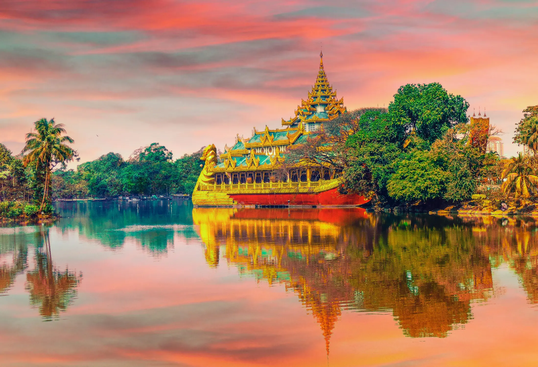 Ornate Burmese royal-style temple or palace structure beside a lake, surrounded by lush tropical trees and reflected in calm water beneath a colorful sunset sky.
