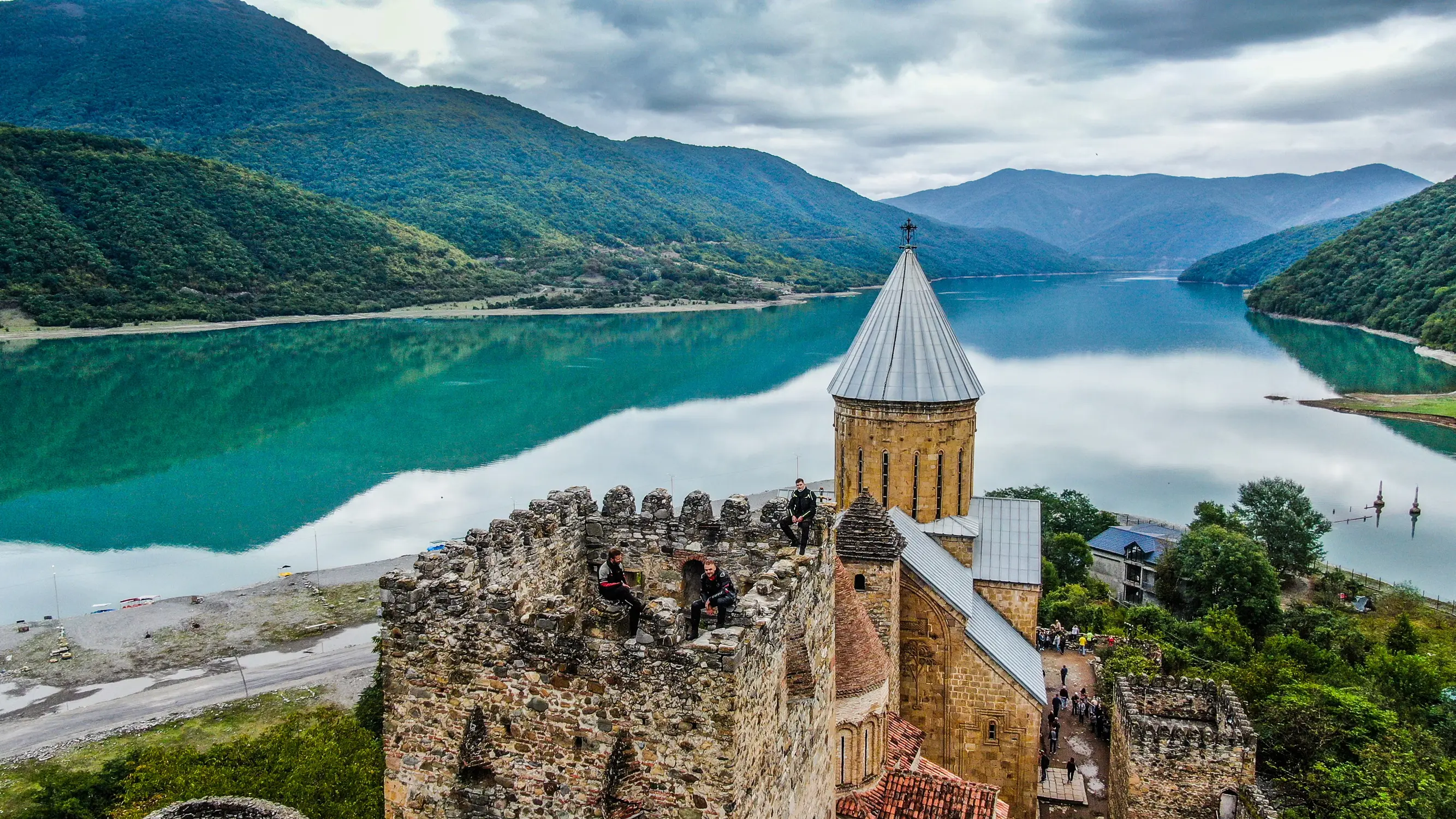 Historic stone church and fortress overlooking a turquoise mountain lake under a cloudy sky.