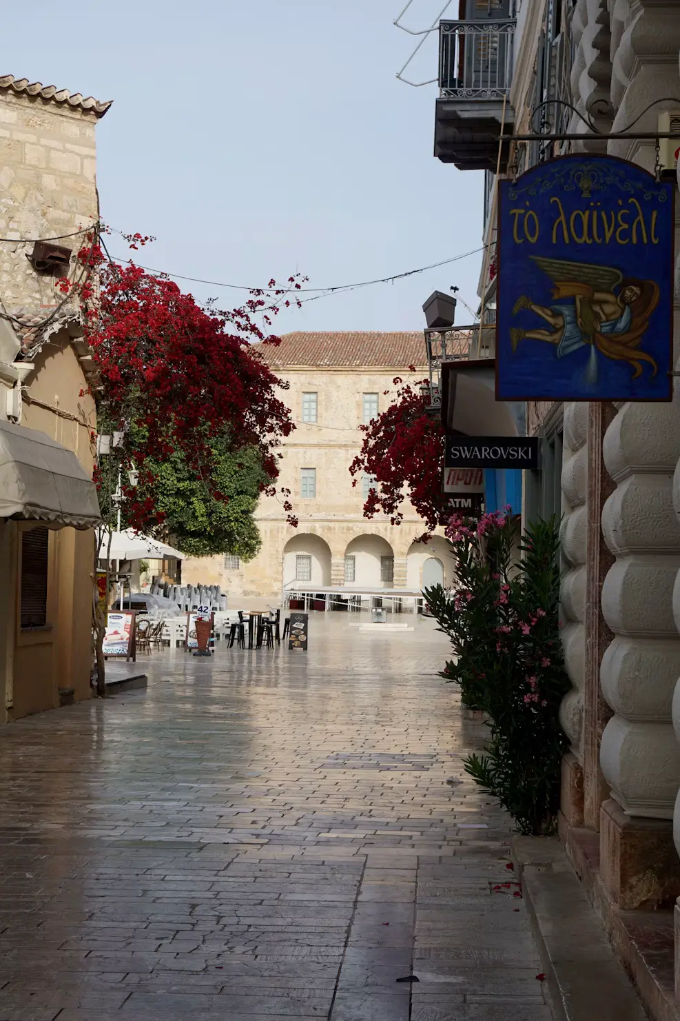 A quiet cobblestone street in a Greek old town, lined with historic buildings and flanked by red flowering plants.