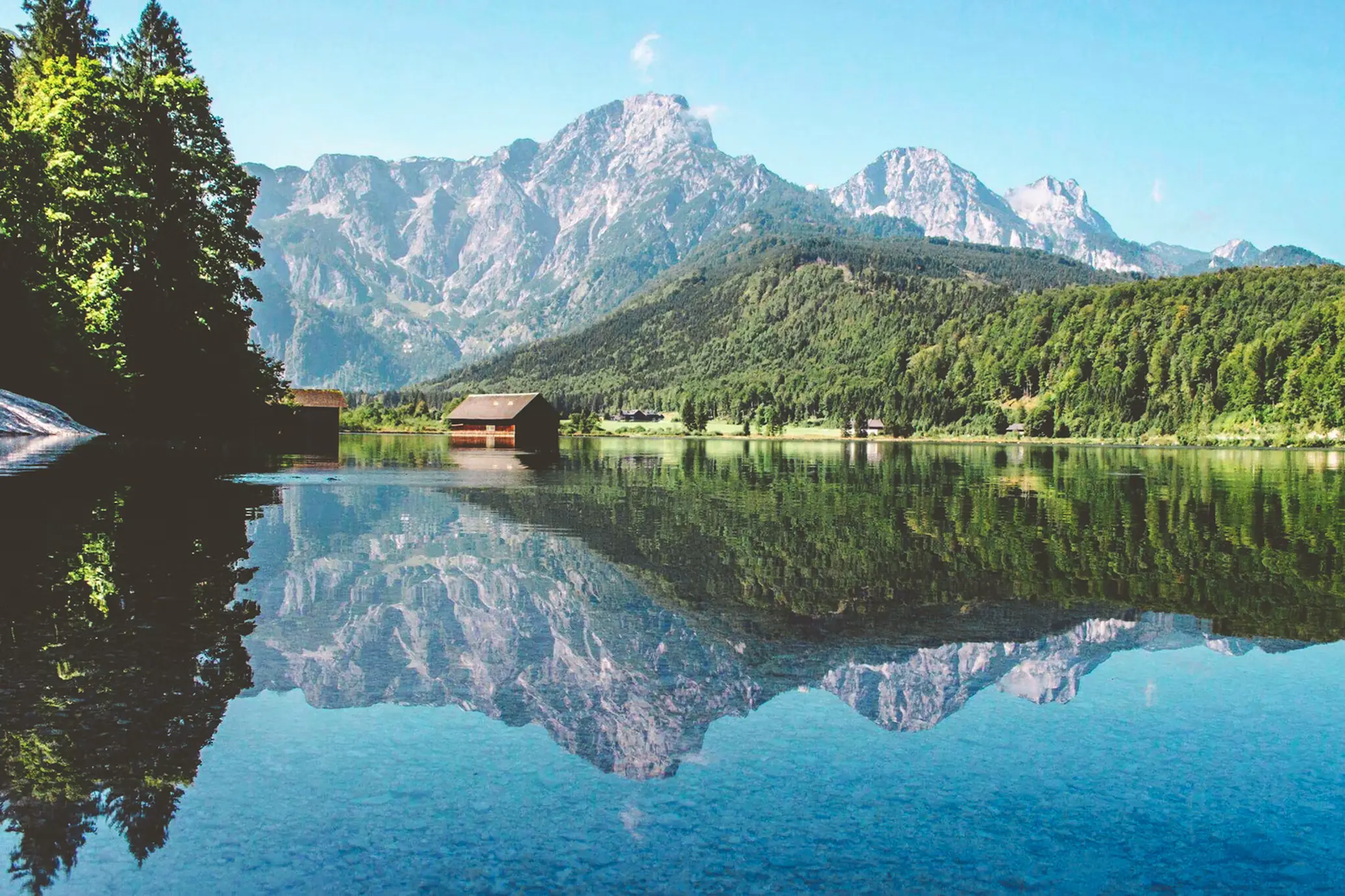 A calm alpine lake perfectly mirrors jagged mountain peaks and a small red boathouse along the tree-lined shore under a clear sky.