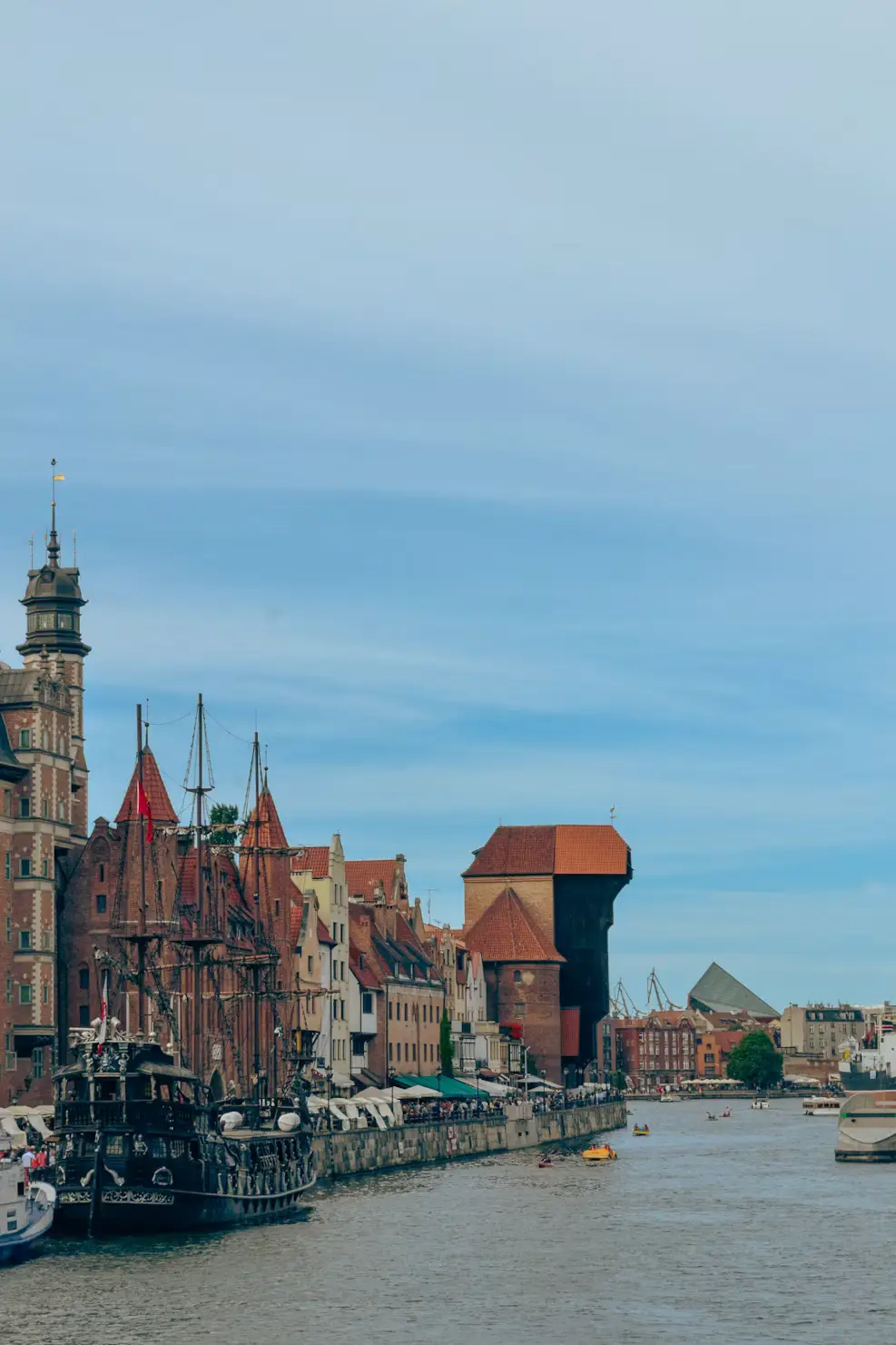 Historic waterfront with traditional European buildings, boats, and a tall church tower under a blue sky.