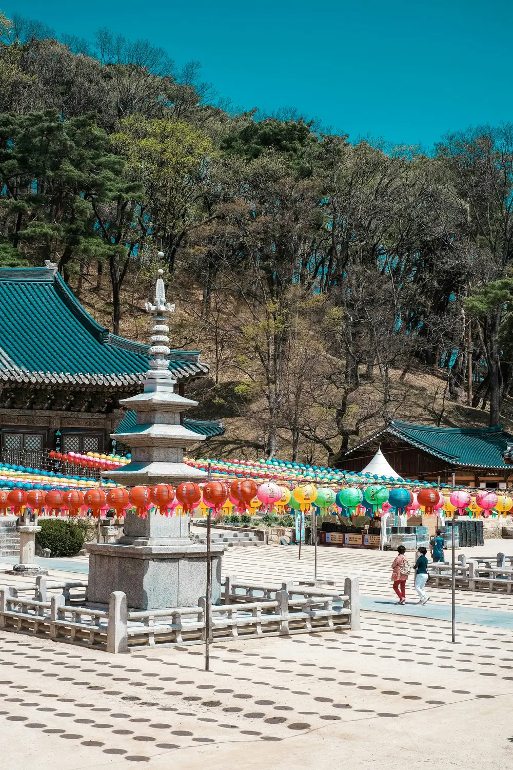 Stone pagoda in a Korean Buddhist temple courtyard decorated with colorful lanterns and surrounded by trees.