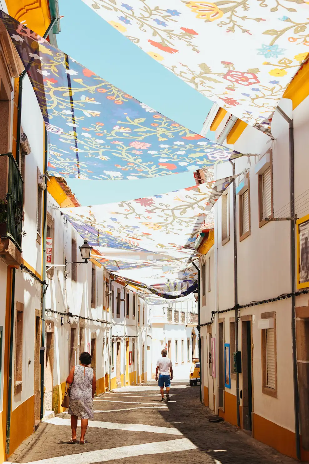 Narrow sunlit street with whitewashed buildings and colorful floral fabric canopies hanging overhead, as a few pedestrians walk along the lane.