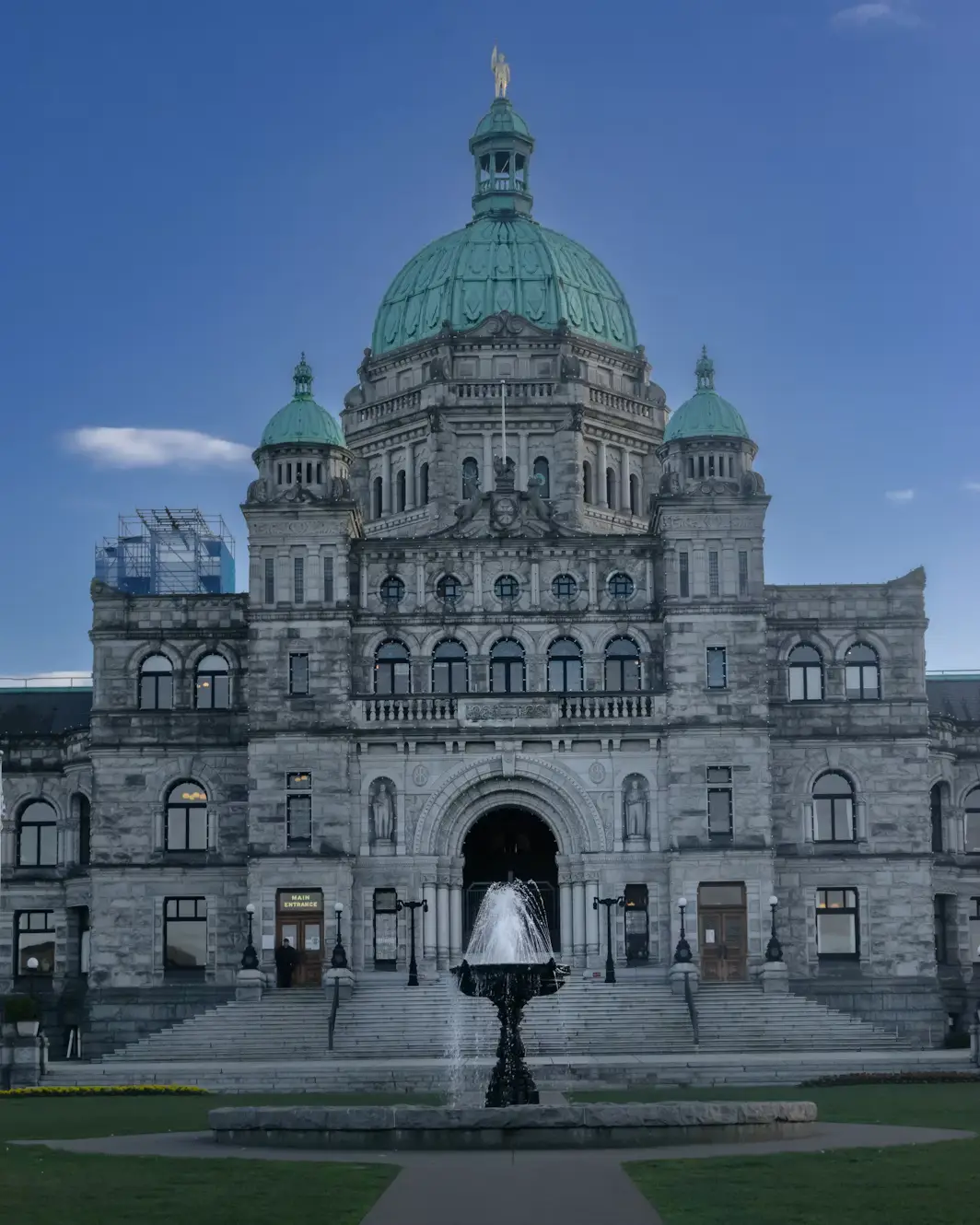 The domed British Columbia Parliament Building in Victoria, with a fountain and manicured lawn in front under a clear blue sky.